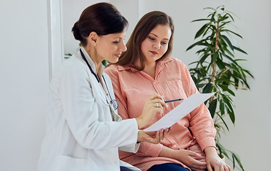 A female physician pointing to a sheet of paper while another woman wearing a peach shirt looks.