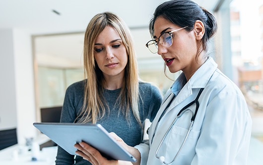 A female physician wearing a white lab coat showing a blonde hair female wearing a green shirt an image on an iPad screen.