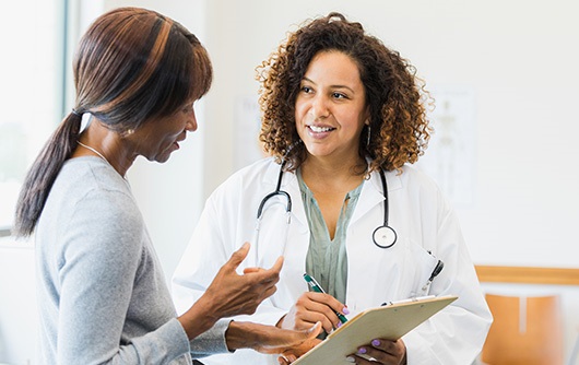 Two women, one a physician wearing a white lab coat with curly brown hair talking in an office.