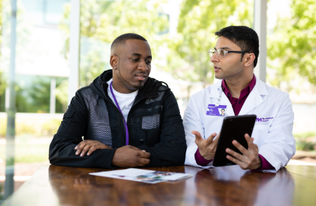 A male Proton physician in a discussion with a young African American male patient while looking at an iPad.