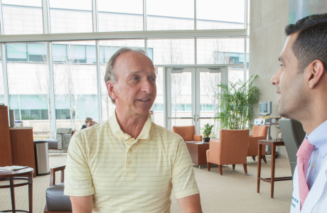 Proton Center physician doctor Mohammed sitting and talking with an older male patient with a yellow striped t-shirt.