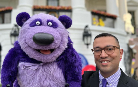 A young man wearing glasses, a collared shirt and purple tie smiling while standing next to a purple Sesame Street puppet.