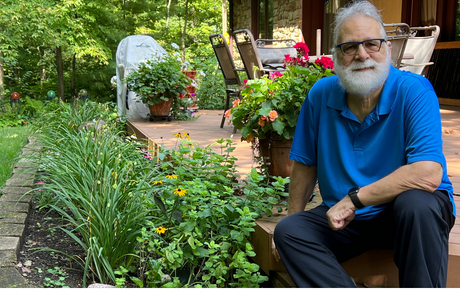 An older man wearing a bright blue shirt sitting next to plants and smiling.