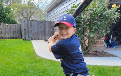 A young boy named Landon smiling at the camera holding his baseball bat and wearing his baseball uniform.