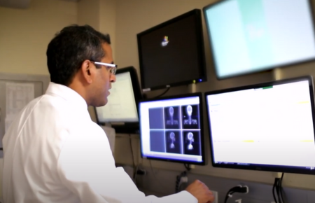 Proton Center physician Doctor Vinai Gondi looking at four computer screens on a wall with various medical images.