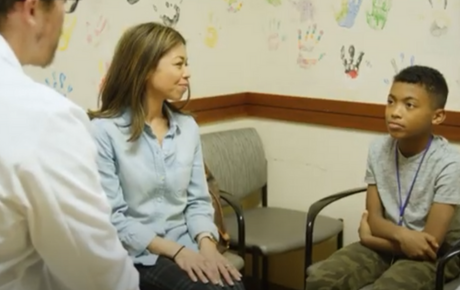 A young male patient sitting in an exam room next to a woman wearing a blue shirt while having a conversation with a male Proton Center doctor.